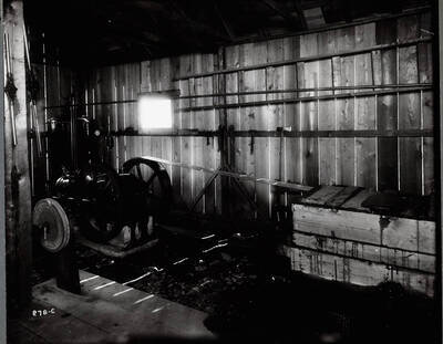 A small air compressor located inside a wooden building. Various tools are hanging on the wall.