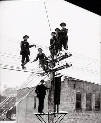 Image shows six telephone men in Wallace, Idaho standing on telephone pole. The building is Masonic Temple (background) and Stevens Meat Market (Foreground).
