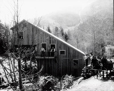 Image of Coeur d'Alene Powder Co., in Wallace, Idaho. Unidentified men are standing around the building, a team of horses are nearby.