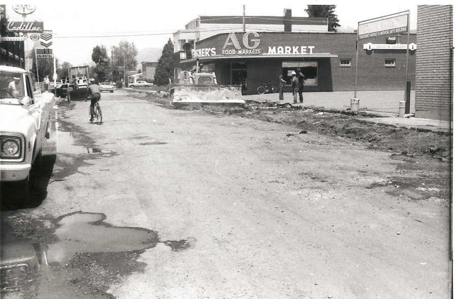 A street scene with a person riding a bicycle on a rough, uneven road. On the left, there is a parked truck and several buildings, including one with a sign reading "Cadillac." On the right, a store with a sign "Gerber's AG Food Markets" is visible, with two people standing near an earth-moving machine. A sign post near the store reads "Thueger's Market & Medical Arts Building" with an additional note "Drive In Grocery."