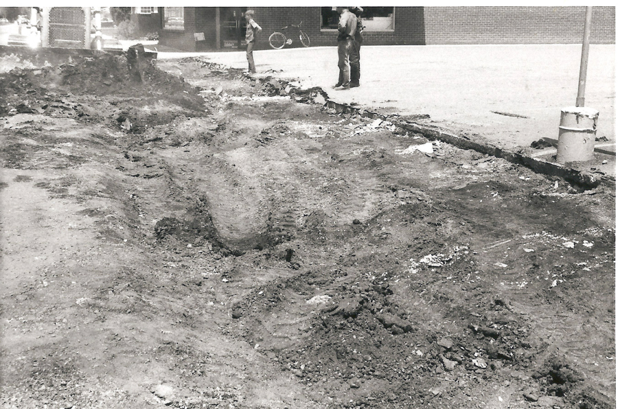 A rough, dug-up street with piles of dirt and debris. Two men stand on the edge of the site, observing the area. A bicycle is leaning against a wall in the background. There's a building with a large window behind them.
