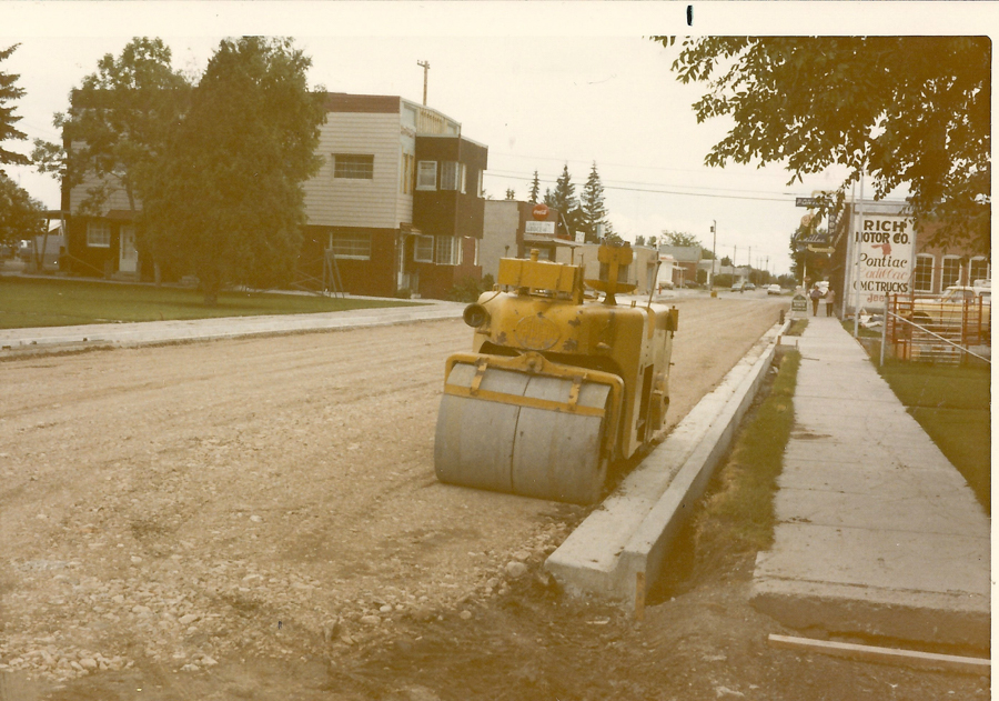 A street scene with a yellow steamroller parked on a dirt road. The sidewalk runs alongside, with trees and buildings lining the street. A sign on one building reads "RICH MOTOR CO. Pontiac Buick GMC TRUCKS Jeep."