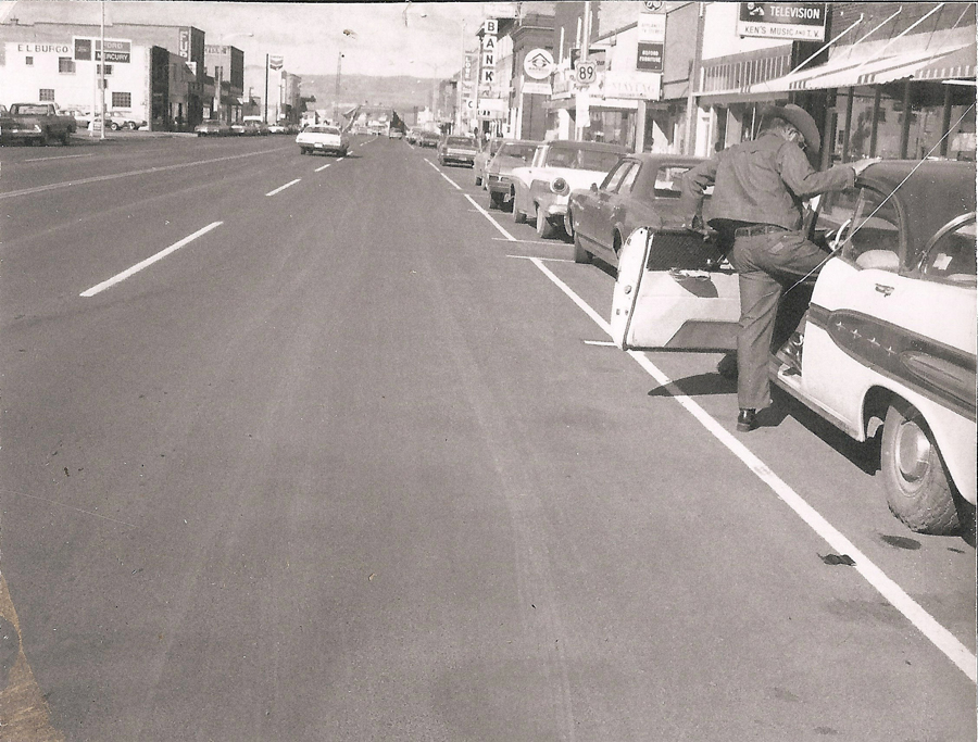 A man in a cowboy hat is stepping into a parked car along a street lined with vehicles. Businesses with signage are visible on the right, including a bank and a television store. The road stretches into the distance with a few cars driving away. There is text for a few businesses, including "EL BUFEO," "BANK," and "TELEVISION."