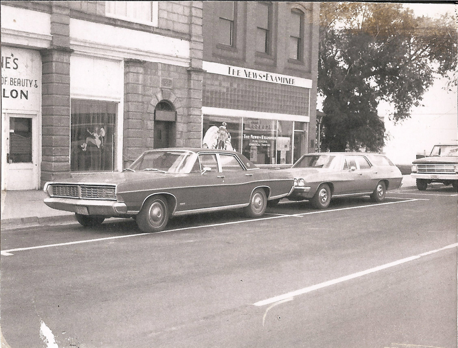 Two vintage cars are parked on a street in front of a building. One car is a sedan and the other is a station wagon. The building has signs that read "The News-Examiner" and "Louise's House of Beauty & Salon." There is a door between the signs and large windows displaying designs or decorations. Trees are visible in the background, alongside another vehicle parked further down the street.
