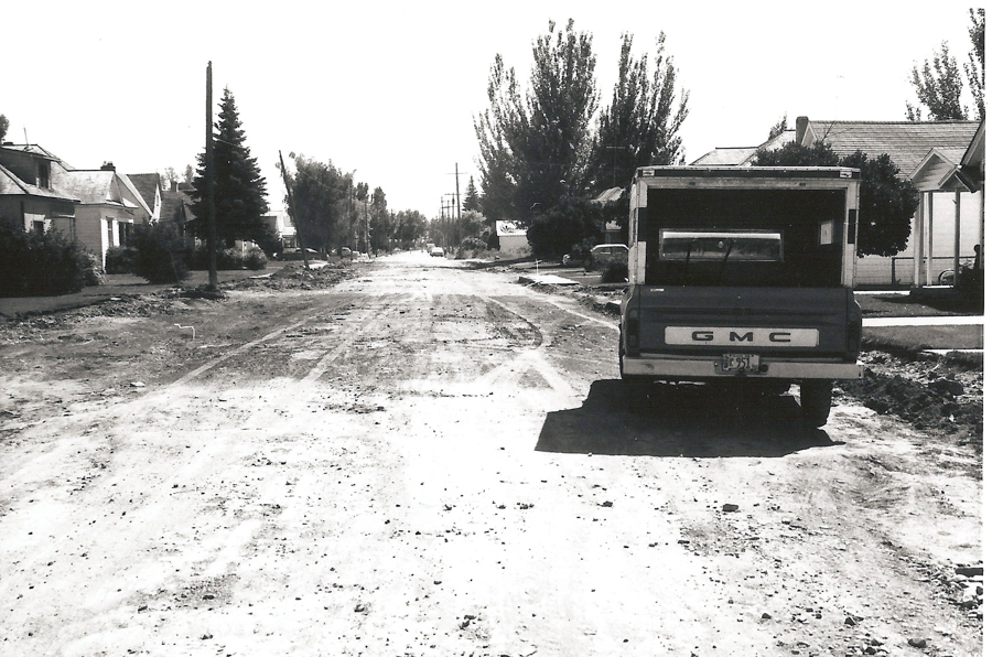 A dirt road lined with houses and trees. A GMC truck is parked on the right side of the road. The road appears uneven with visible tracks and debris.