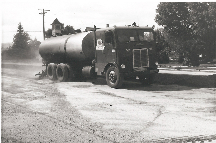 A large tanker truck labeled with the number "910" is parked on a road. There is a logo with a sun design on the side of the cab. In the background, there are trees, a telephone pole, and a building with a pointed roof. The road appears dusty.