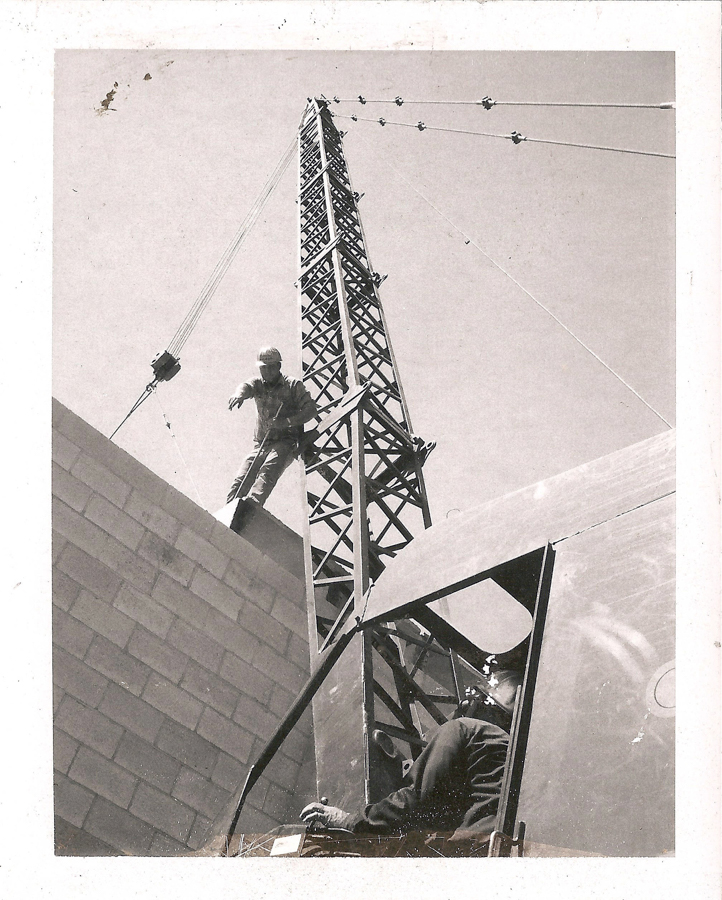 A person standing on a steel structure at a construction site, wearing work clothes and a hat. Another person is seated inside a control cabin at the base, operating machinery. The scene includes a tall metal framework and a partially constructed brick wall.