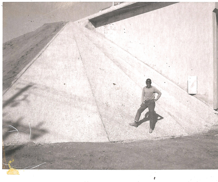 A person stands on a sloping concrete surface, leaning slightly with one hand on their hip. Behind them is a steep, triangular structure adjacent to a building wall. The ground in front is dirt, and some foliage appears in the bottom left corner.