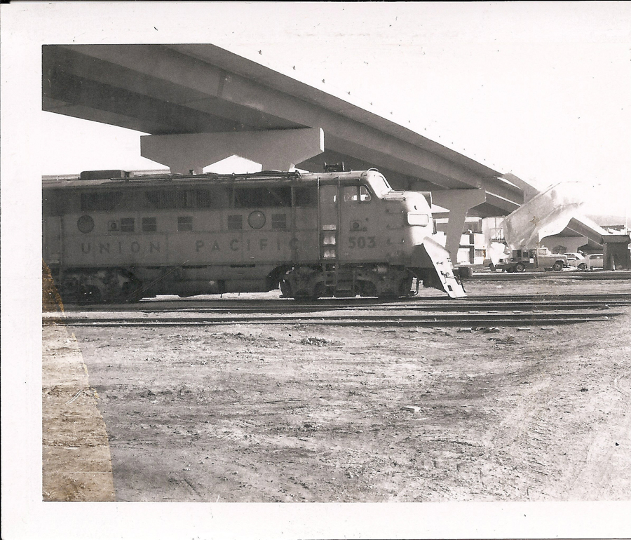 A train engine labeled "Union Pacific 503" is on railway tracks. There is a large, elevated highway structure in the background with a vehicle parked beneath it. The area appears to be an industrial or construction site with dirt ground and some scattered equipment.