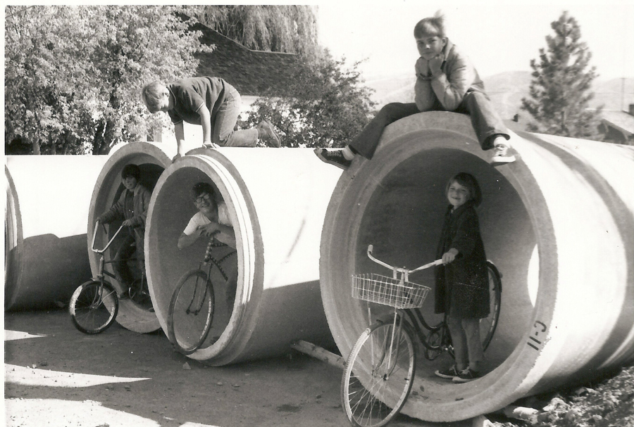 Four children are playing with bicycles around large concrete pipes. Two children are inside the pipes with bikes, one is climbing over the top, and another is sitting on top of one of the pipes, resting their chin on their hands. Trees and a building can be seen in the background.