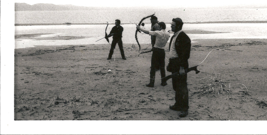 Three people standing on a sandy beach by the water, each holding a bow and aiming it. The figure in the middle draws the bowstring back, preparing to shoot. The background features distant hills or mountains across the water. Sparse vegetation and sticks are scattered on the sand.