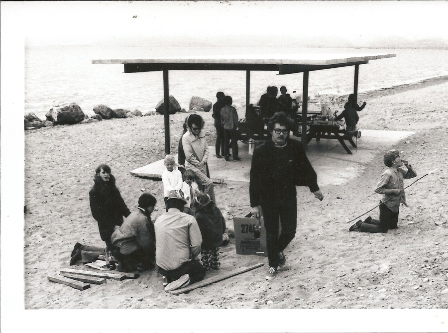 A group of people gathered on a sandy beach by the water, with a covered picnic area in the background. Several adults and children are sitting, kneeling, and working with wooden planks on the sand. A person is walking, carrying a box labeled "Z714" with some text underneath. A few individuals are at picnic tables, and a child is playing nearby with a stick. The scene conveys a casual, communal beach gathering.