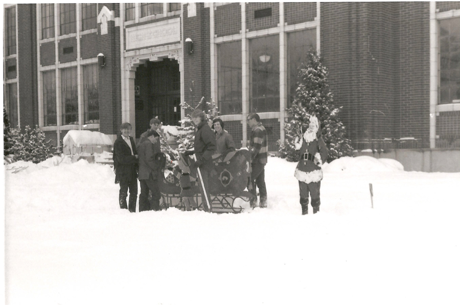 A group of people standing in the snow near a sleigh, in front of a building with large windows. One person is dressed as Santa Claus. There are evergreen trees covered in snow, and the building has a sign above the entrance that is difficult to read.