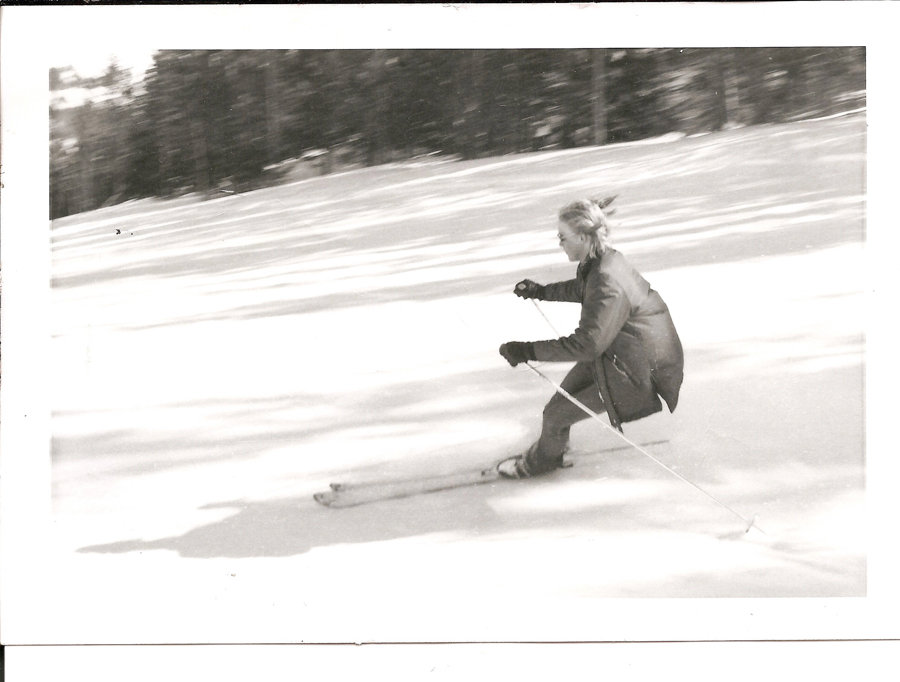 A person skiing quickly down a snowy slope, wearing a jacket and gloves, with trees visible in the background.