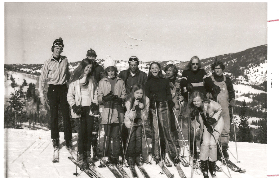 A group of people is standing on a snowy mountain slope. They are wearing winter clothing and holding ski poles. The background shows snow-covered hills and trees.