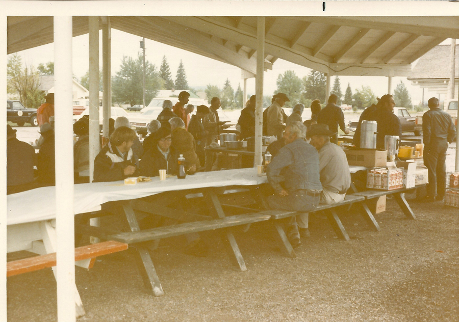 People are gathered under a pavilion. Some are sitting at picnic tables covered with white paper, eating and talking. A few are wearing hats and casual clothing. In the background, others are standing near tables with various food items and large drink dispensers. Cars and trees are visible in the open area beyond the pavilion.