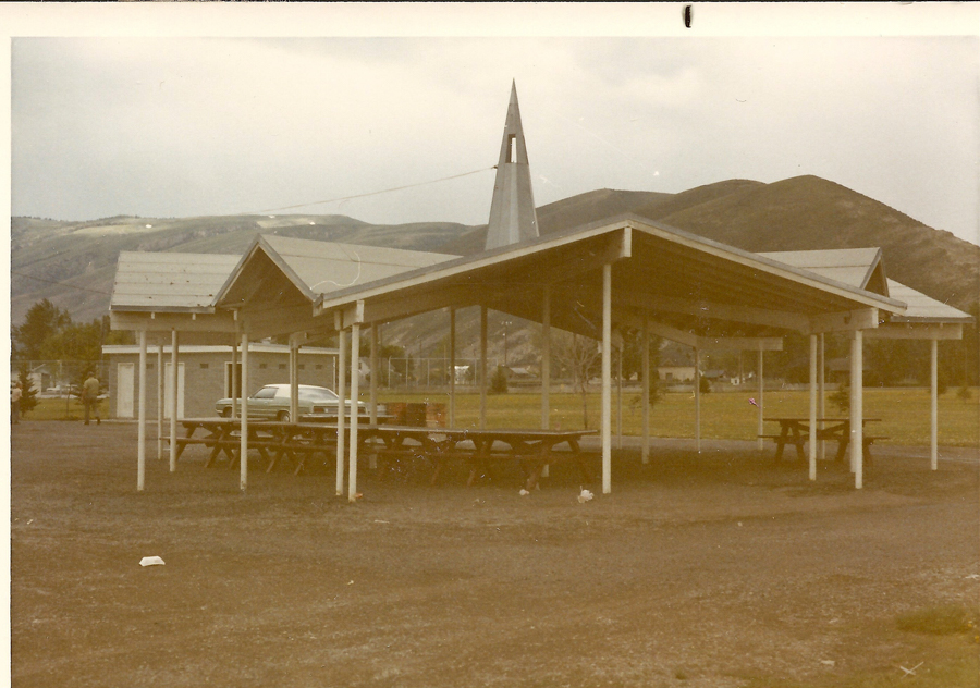 A covered picnic area with several picnic tables underneath. In the background, there are mountains and a pointed structure resembling a steeple. A car is parked nearby, and a person is walking towards a small building. The scene is set in an open field.