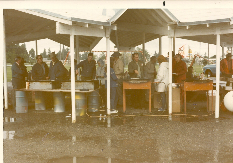 A group of people, including men and women, are gathered under a covered pavilion. They appear to be cooking or preparing food on large grills set on barrels and tables. There is a wet ground with some puddles. In the background, there is a sign that includes the word "FOR SALE" and a playground with swings. A blue vehicle is partially visible.