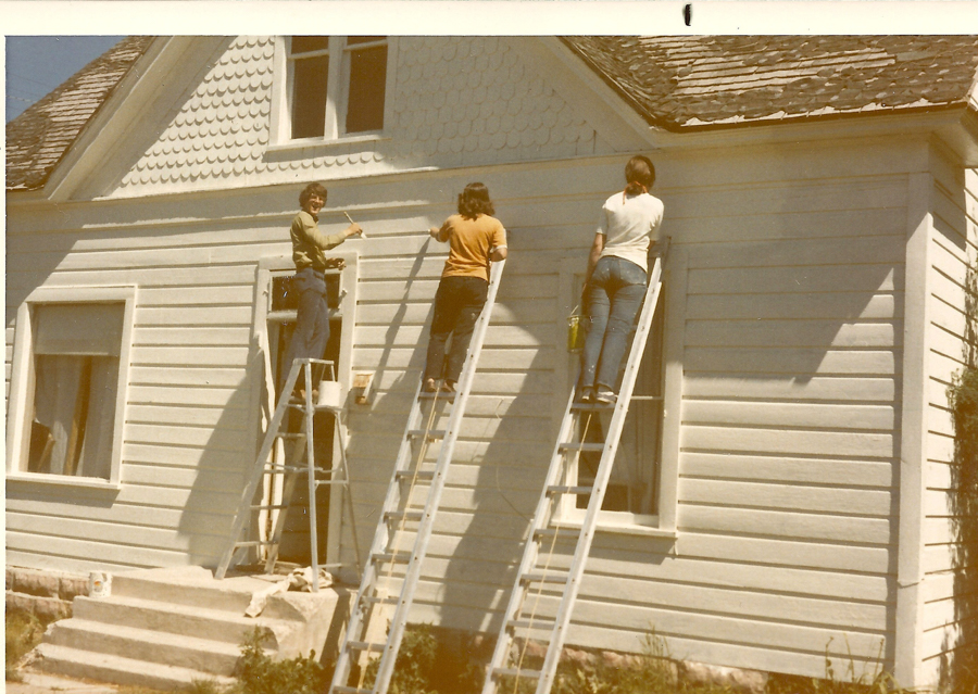 Three people are painting the exterior wall of a house while standing on ladders. One person is on the left ladder, holding a paintbrush and smiling. The second person is in the middle, painting with a brush. The third person on the right ladder holds a bucket. The house has overlapping wooden siding, a shingled roof, and features multiple windows.