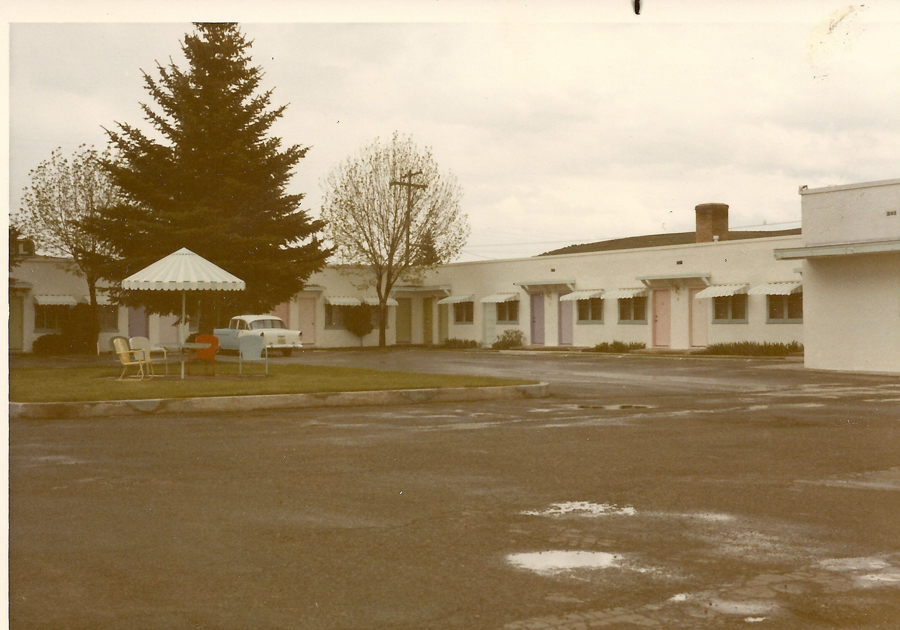 A row of single-story motel rooms, each with a different colored door and a small awning above the windows, is shown along with a parking area. In the foreground, there is an arrangement of outdoor furniture including a table and several chairs under a striped umbrella. A large tree and a car are visible near the building. The area appears to be wet, suggesting recent rain.