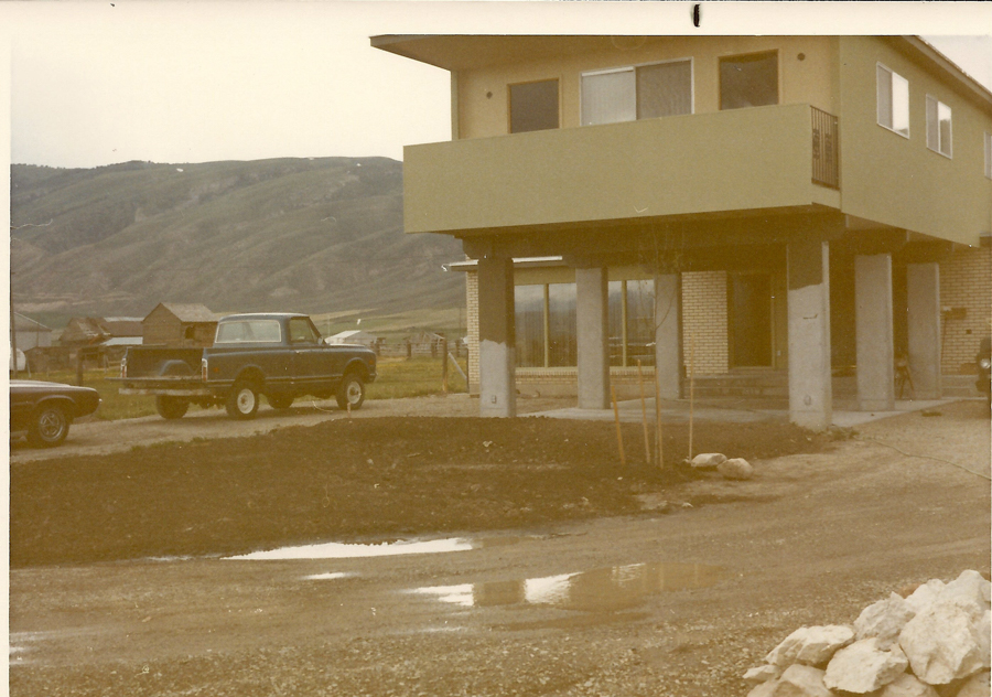 A two-story house with a prominent balcony on the upper level, supported by columns. There is a dirt driveway in front, with a pickup truck parked alongside a car. The background shows a rural landscape with hills and a few small buildings.
