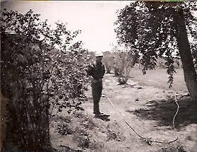 A person standing in an outdoor area with trees and bushes, holding a hose. The ground appears to be grassy with some patches of dirt. There are larger trees on the right side and smaller shrubs on the left. The scene looks open and spacious.