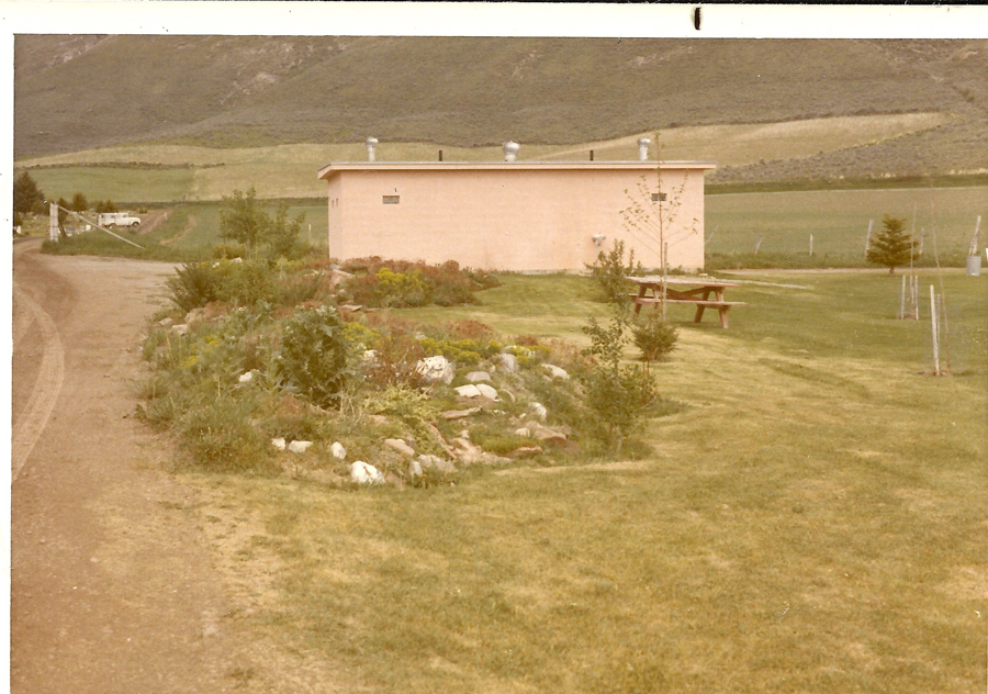 A small rectangular building with several vents on the roof is situated in a grassy area. In the foreground is a landscaped garden with various plants and rocks. To the right, a picnic table is set on the lawn. A dirt road runs alongside the garden, leading towards a distant white vehicle. In the background, rolling hills and fields stretch into the distance.