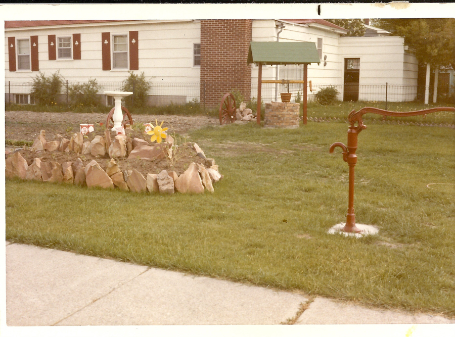 A yard with a red hand pump standing on grassy ground. In the background, there is a circular garden bed edged with large stones, containing a small white birdbath, decorative flowers, and small objects. A small brick well with a green roof is to the right of this garden bed. A house with red shutters and a brick chimney forms the backdrop.