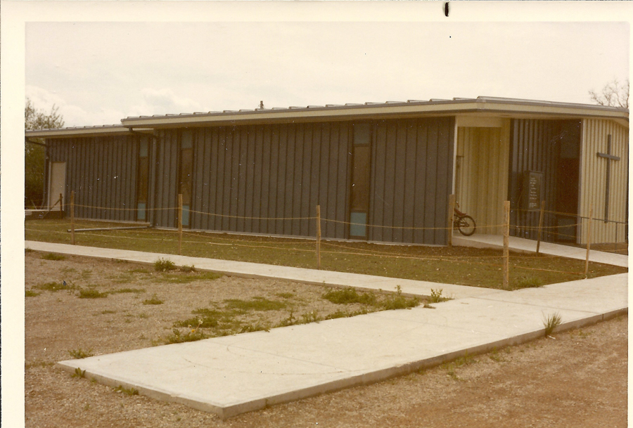 A one-story building with vertical siding and multiple rectangular windows. A bike is parked near the entrance, which has a covered area. A sign with unreadable text is displayed near the entrance. A paved walkway leads to the building, bordered by grass and dirt areas with some sparse vegetation, enclosed by a rope fence.