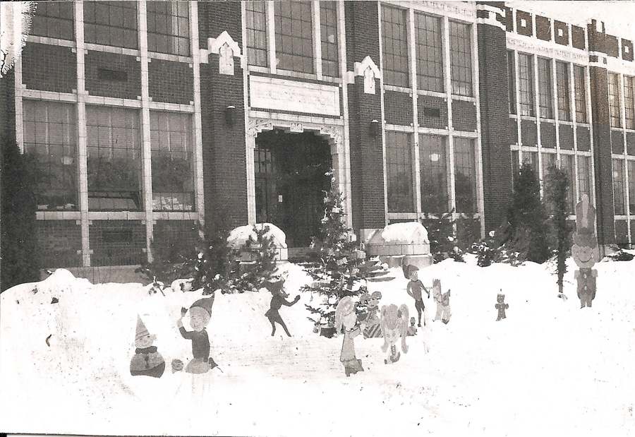 A large brick building with multiple tall windows in the background. In the foreground, there is snow on the ground and several cut-out figures of children and animals scattered throughout, suggesting a festive or playful winter scene. Small evergreen trees are positioned along the entrance steps.