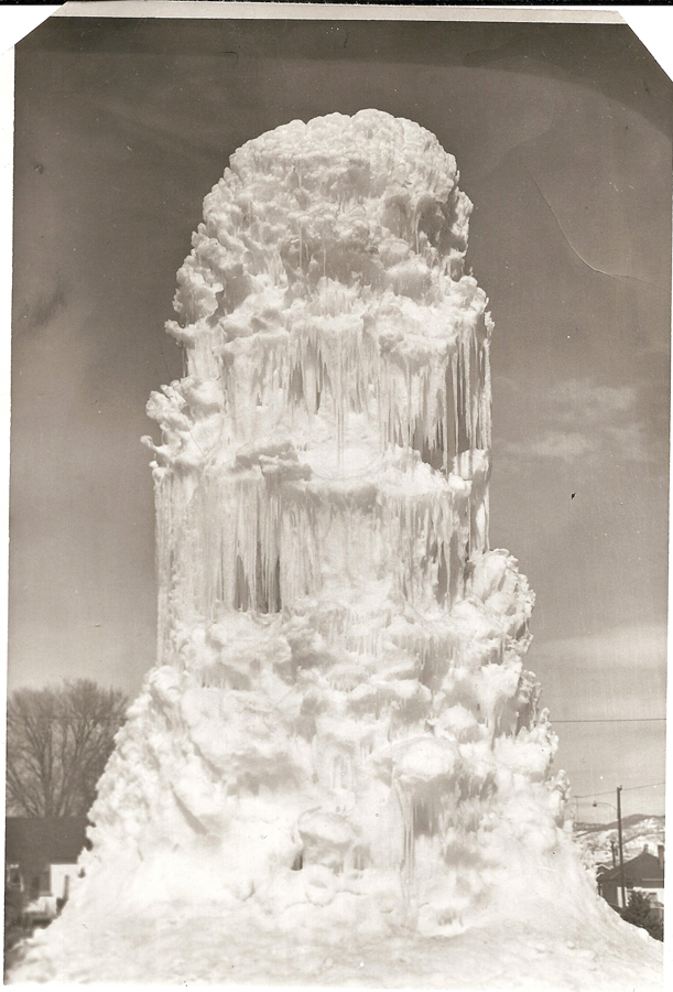 A large, natural ice formation resembling a towering column is prominently displayed. The structure is covered with icicles and snow, and trees and buildings are visible in the background.