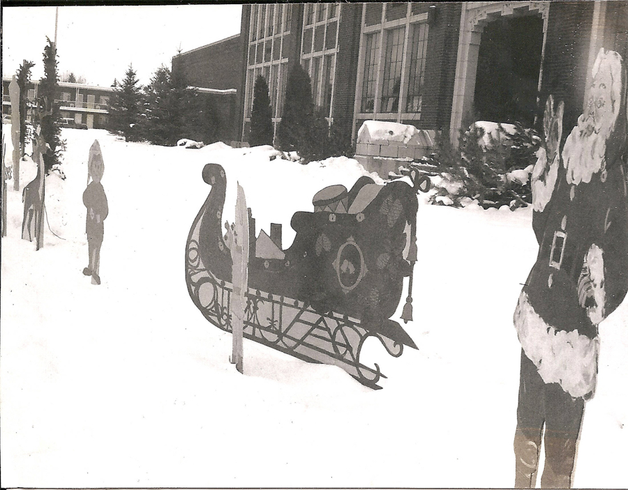 A snowy outdoor scene featuring a decorative sleigh surrounded by cutouts of festive characters. In the background, there are large windows and a building with shrubs covered in snow.