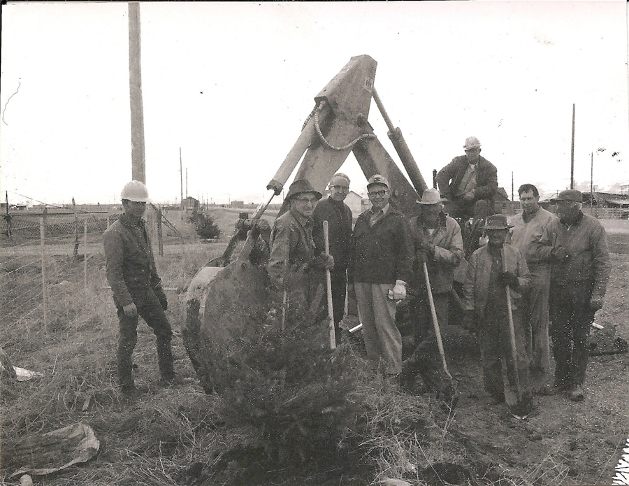 A group of men stand around a large piece of construction equipment in an open field. Some are holding shovels, and a few are wearing hard hats. A small tree is in the foreground. There are utility poles and a fence in the background.