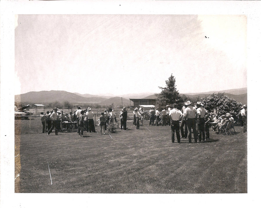 A group of people gathered on a grassy area, with mountains visible in the background. Some individuals are standing, while others are seated on chairs. A few people wear cowboy hats. There is a tree and a bush, as well as a small building and an aircraft in the distance.