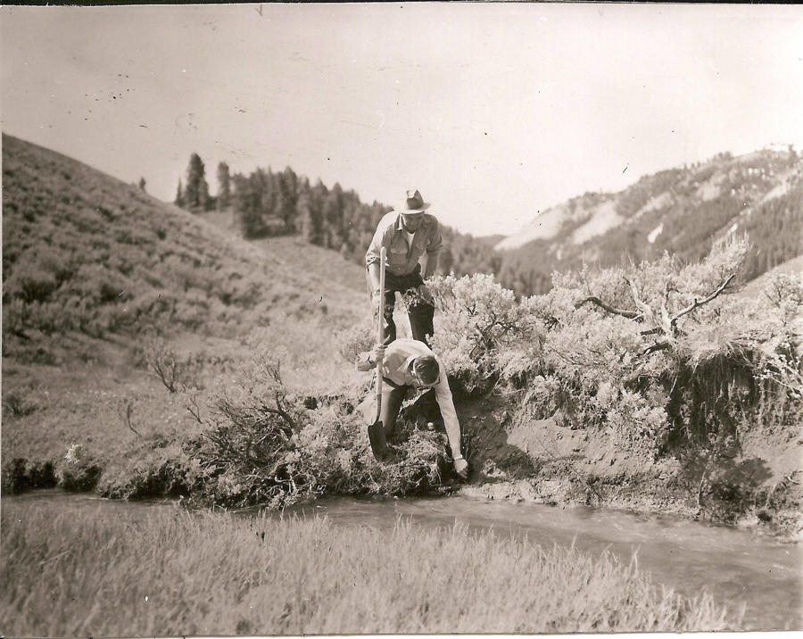 Two men are in a natural setting with hills and trees in the background. One man is bent over next to a small stream, and the other man stands beside him, leaning forward slightly. There is vegetation around them, and the landscape is rural and somewhat rugged.