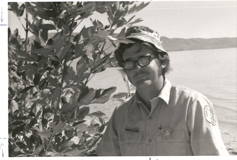 A person wearing a hat and glasses stands next to a leafy bush near a body of water. They are wearing a shirt with a patch on the sleeve that reads "Idaho State Parks." A badge on their shirt says "Park Ranger." Hills are visible in the background.