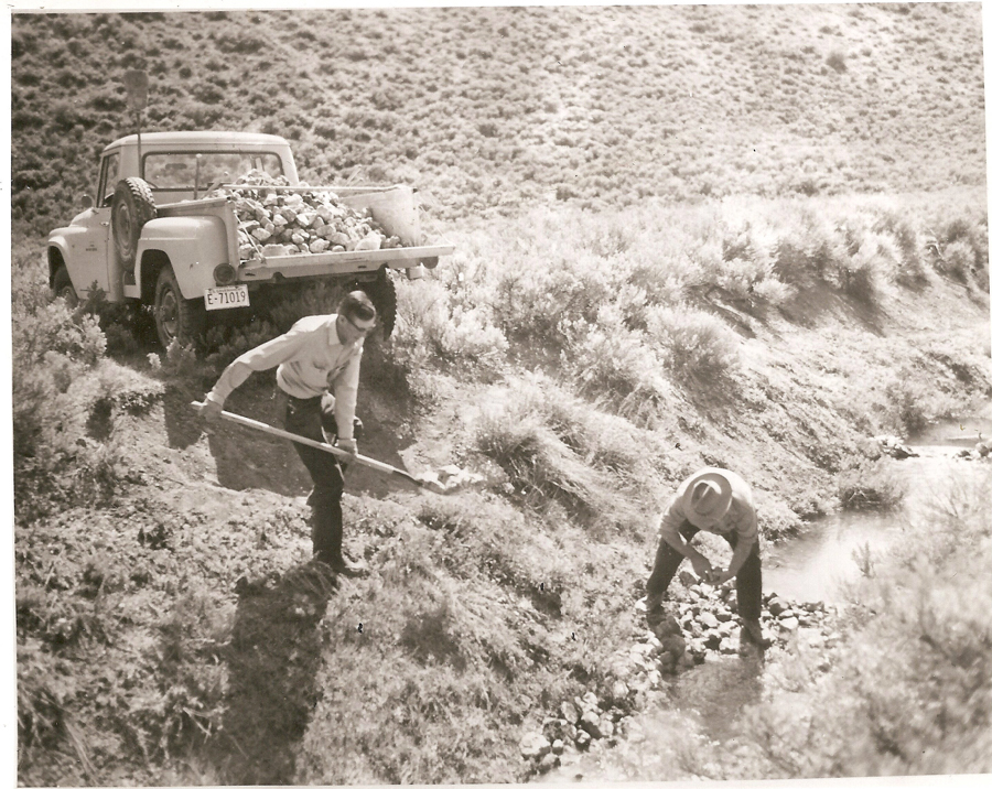 Two people working near a stream in a rural area, one using a shovel and the other picking up rocks. A truck loaded with rocks is parked nearby. The terrain is hilly with scrub vegetation. The truck's license plate reads "E 71019."