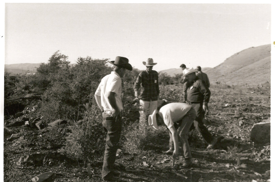 A group of people wearing hats standing outdoors on a hillside. One person is bending over, examining something on the ground. The landscape includes bushes and distant hills.