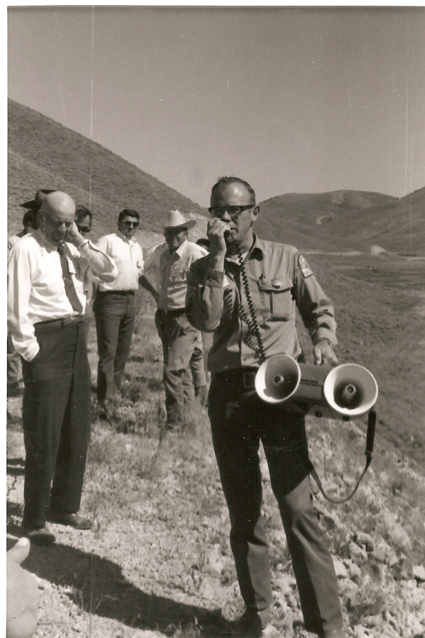 A group of men standing on a hillside. One man in the foreground is holding a megaphone and speaking into a microphone. He is wearing glasses and a uniform with patches on the sleeves. The others are dressed in shirts and ties or hats, standing slightly behind him, looking in different directions. The background consists of rolling hills.