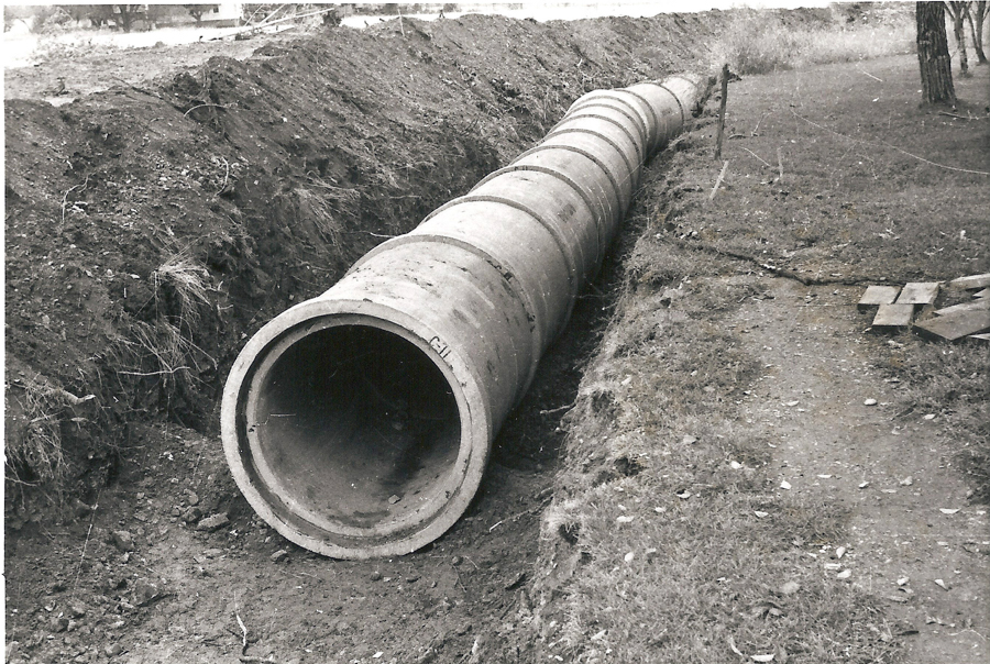 A large concrete pipe laying in a trench, surrounded by earth and grass. A path runs alongside the trench. There are wooden planks and a tree visible on the right side.