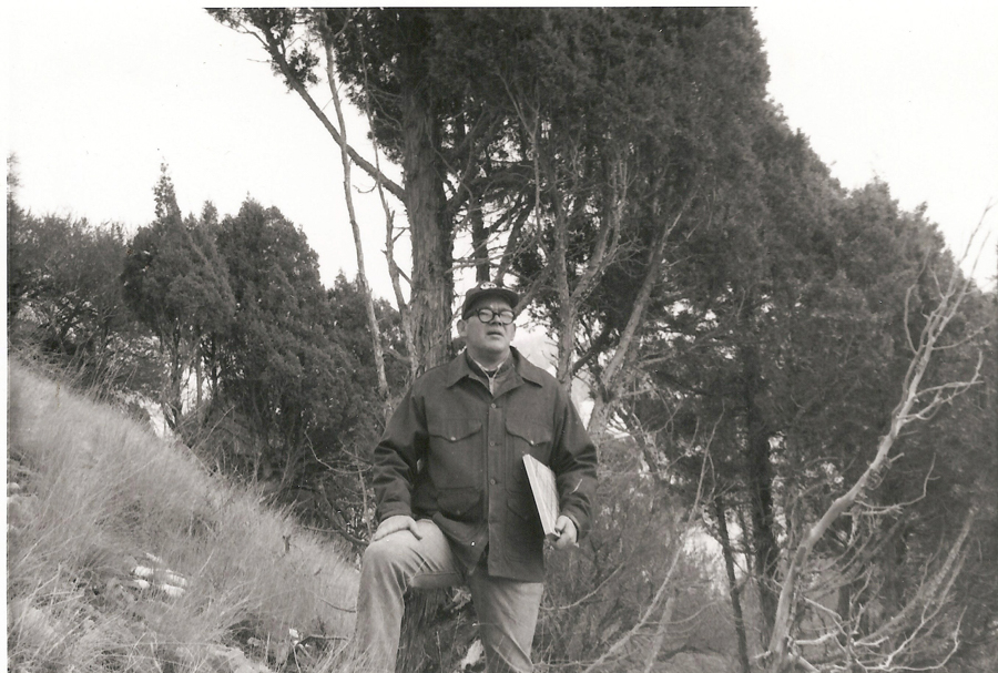 A man wearing a jacket and cap stands outdoors with one foot resting on a rock. He holds a folder in one hand and is surrounded by tall trees and grass.