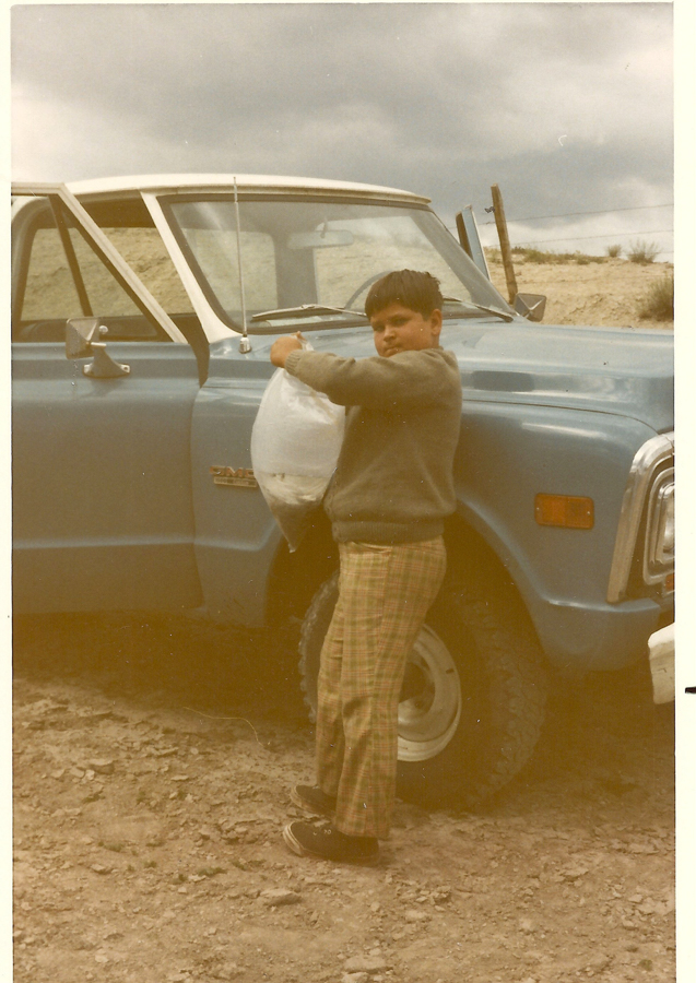 A young boy standing next to a blue truck, holding a plastic bag. He is wearing a sweater and plaid pants. The truck has the word "Custom" visible on the side. The scene is set outdoors on a cloudy day.