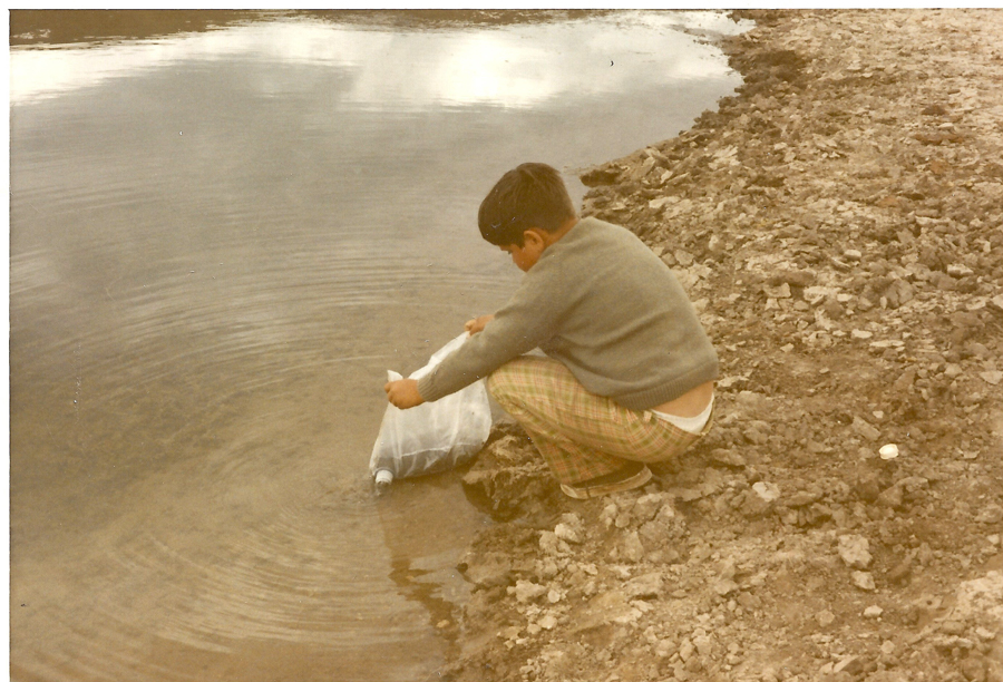 A child wearing a sweater and plaid pants is crouching by the edge of a body of water, dipping a clear plastic bag into the water. The ground near the child is rocky and uneven.