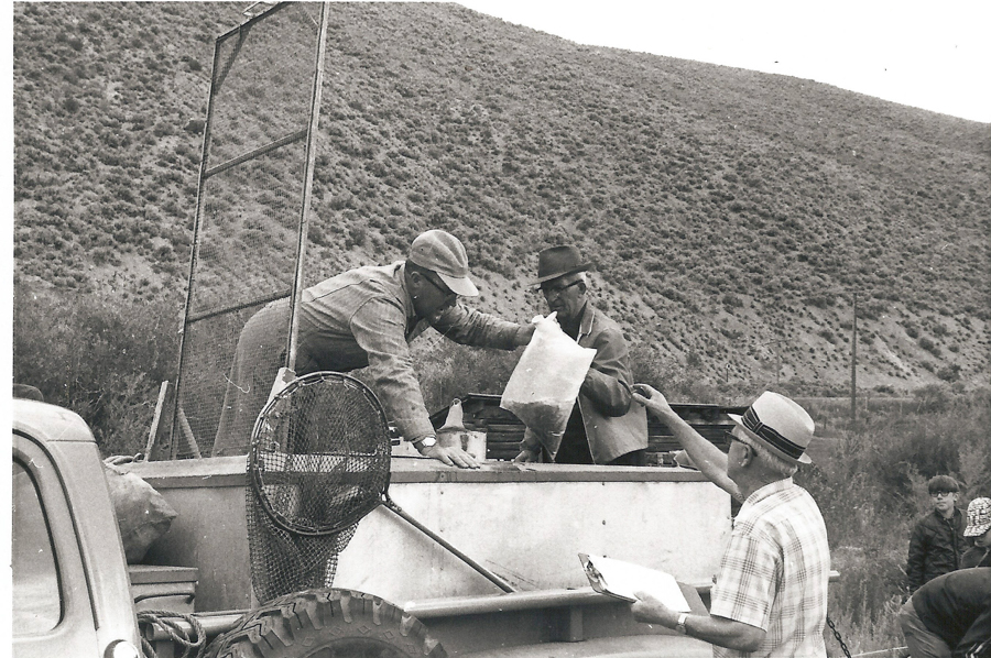 Three men are standing around a pickup truck near a grassy hillside. One man is handing a large bag to another man who is in the truck bed. The third man is holding a clipboard and appears to be writing something. A large net is attached to the side of the truck. Two additional people are partially visible in the background.