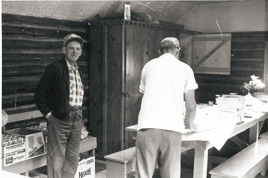 A man is standing with his hands in his pockets, wearing a plaid shirt, jacket, and cap. To his left are boxes labeled "Dr Pepper," "Fanta," and "HEATH." Another man is facing away, leaning over a table covered with a tablecloth and various packaged items. The room has wooden walls and a cabinet in the background.