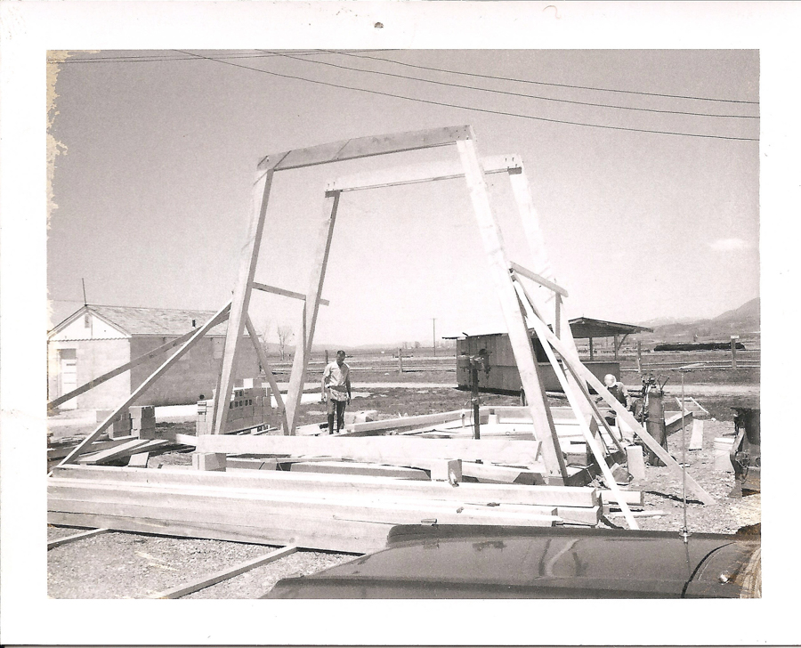 Several people working on a large wooden structure under construction. A parked car is visible in the foreground, and in the background are utility lines, a small building, and open fields with mountains in the distance.