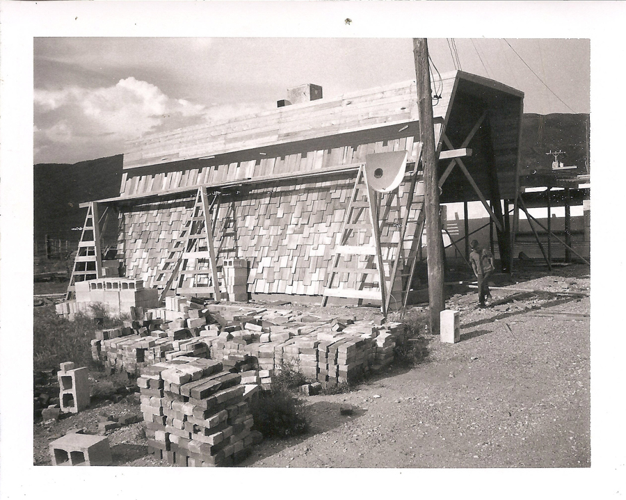 A man standing near a partially constructed building with a wooden exterior covered in shingles. There are two ladders leaning against the structure. Several stacks of bricks and concrete blocks are scattered on the ground around the site. A utility pole is nearby, and the background shows some hills under a cloudy sky.