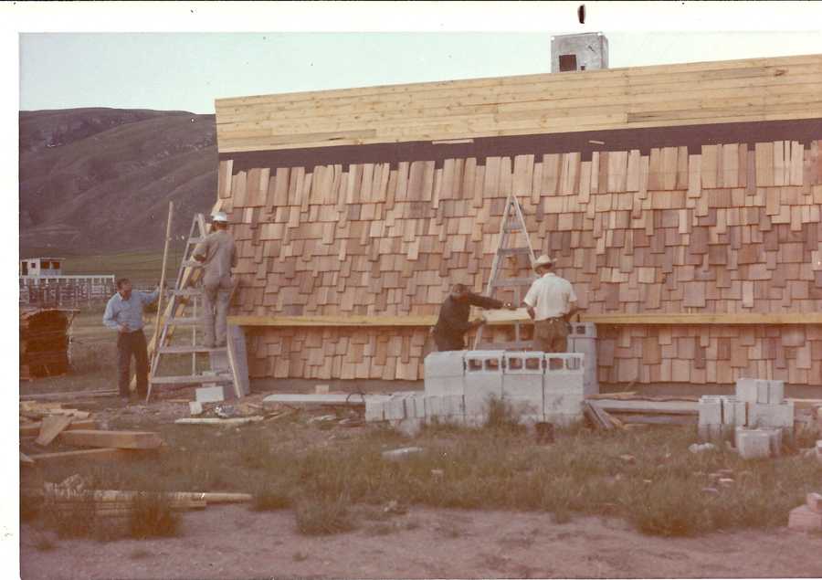 Four people are working on the construction of a building covered with wooden shingles. Two individuals are on ladders, while another is bending over near a stack of concrete blocks. The scene is outdoors in a grassy area with hills in the background. Building materials are scattered on the ground.