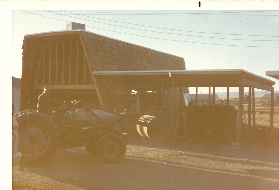 A person sitting on a tractor in front of a building with a slanted roof, featuring wooden shingles and an open upper section. A covered area extends from the building, with hills and open fields visible in the background. Power lines are seen overhead.