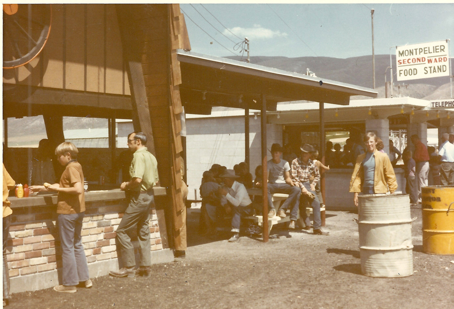 A group of people is gathered around a food stand with a brick façade. Some are standing and others are seated at picnic tables under a shelter. On the structure behind them is a sign that reads "Montpelier Second Ward Food Stand." In the foreground, there is a barrel, and a telephone booth sign is visible in the background. A few people are wearing cowboy hats and casual clothing.